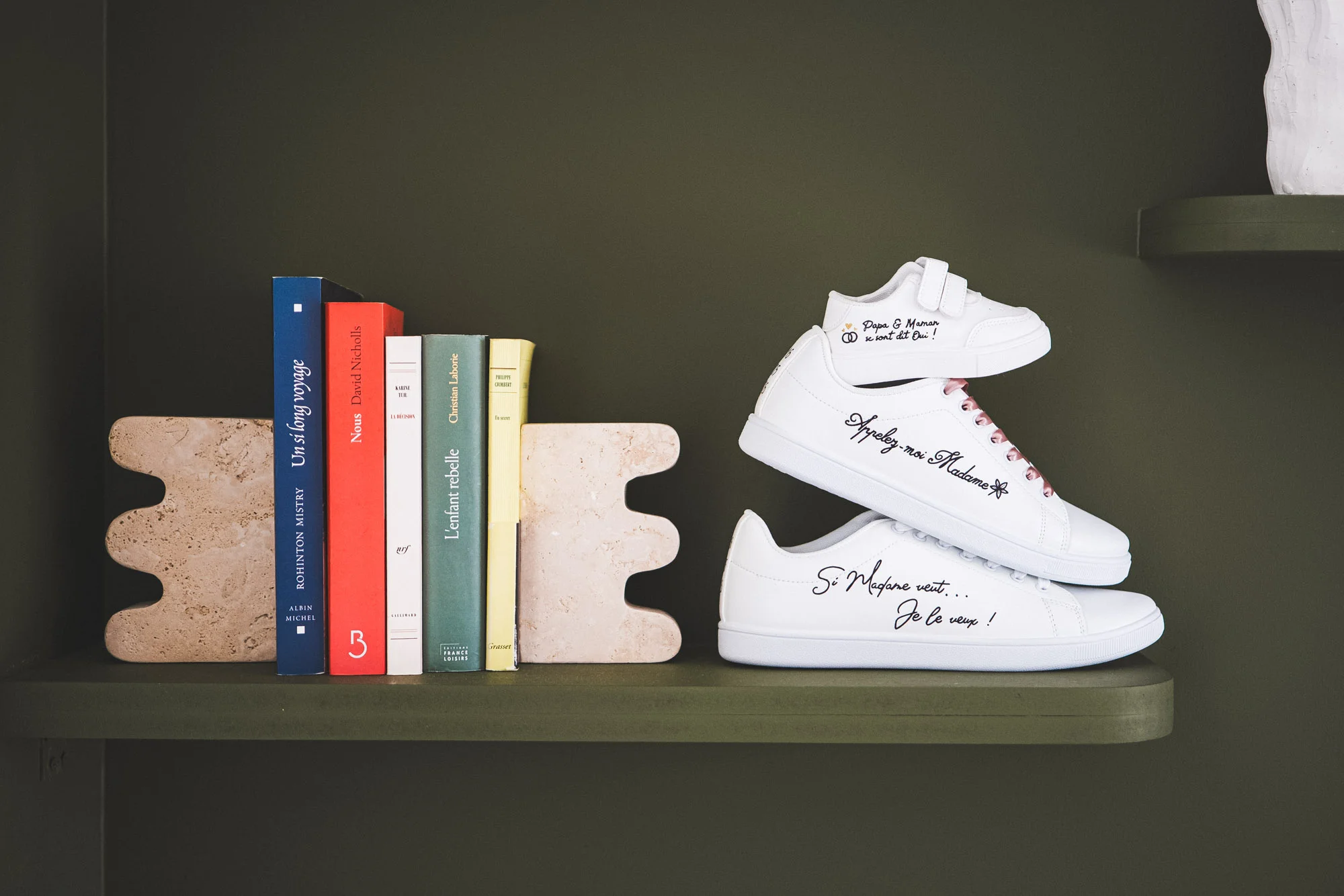 Chaussures de mariage blanches personnalisées posées sur une étagère près de livres colorés.