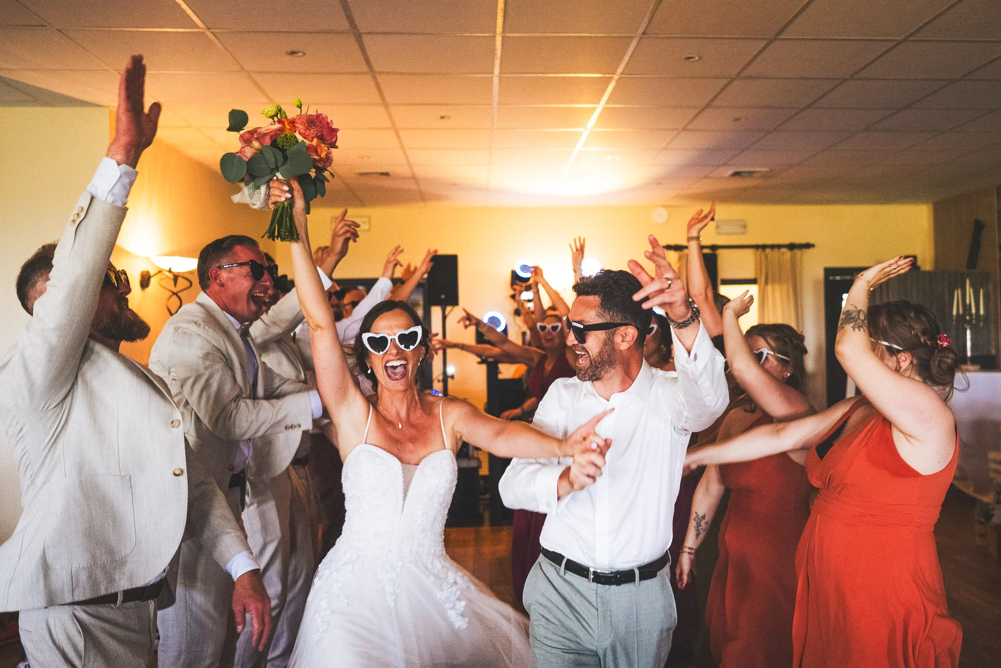 Mariée levant son bouquet au milieu des invités qui dansent avec des lunettes fantaisie.
