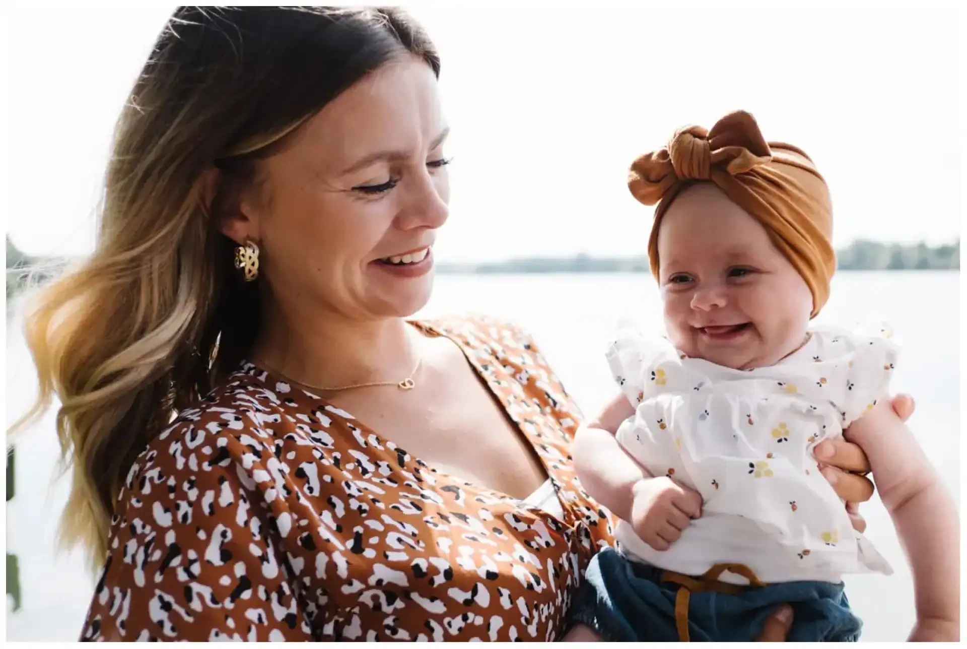 Une mère souriante tenant son bébé coiffé d'un bandeau brun au bord de l'eau.