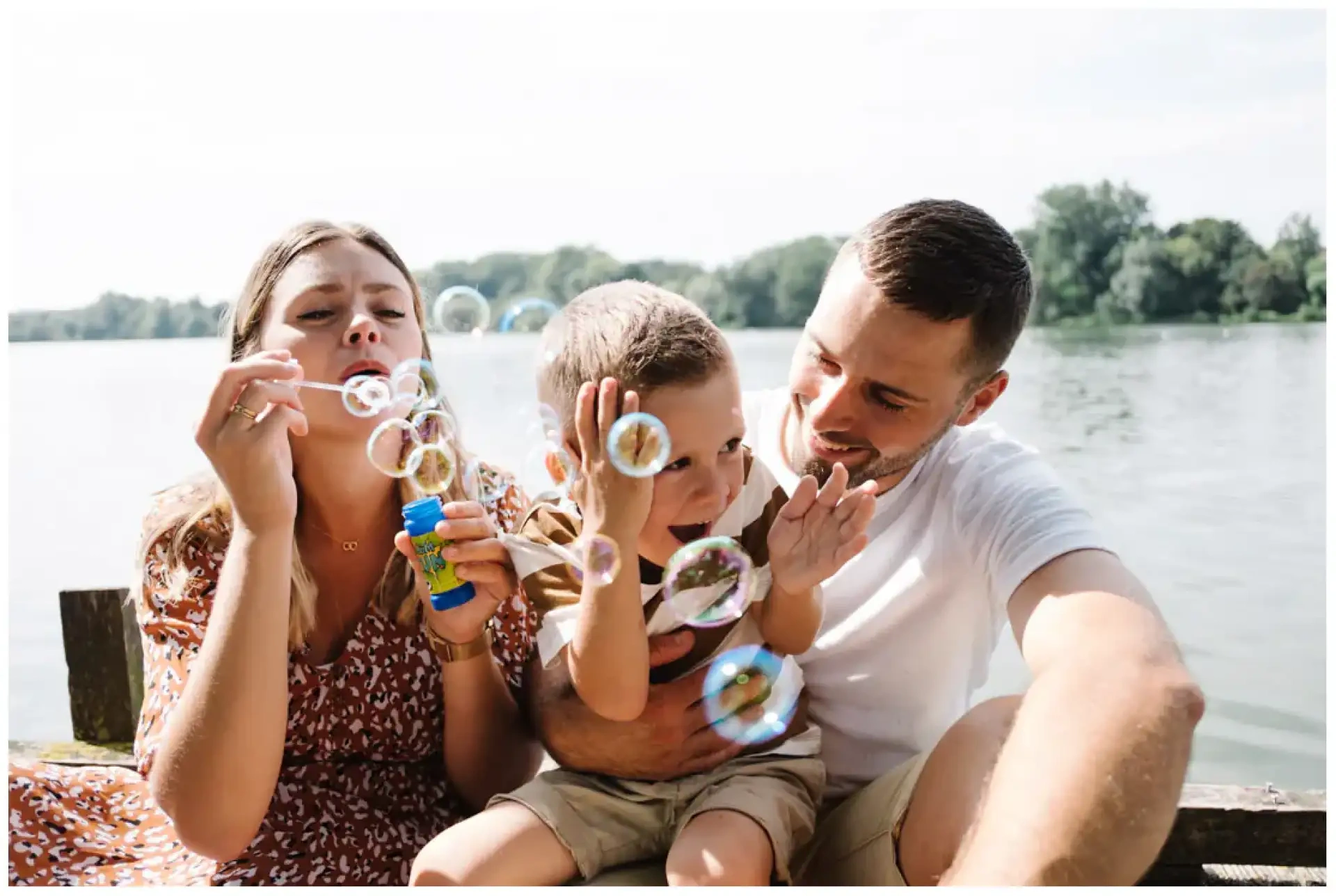 Deux parents et leur jeune garçon jouant avec des bulles de savon au bord d'un plan d'eau.